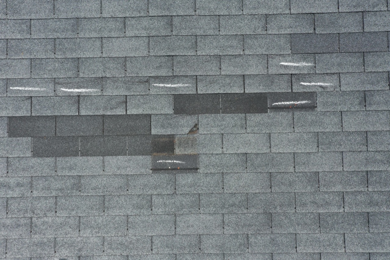 Lifted and creased shingle tabs marked in chalk during a roof inspection, beside darker patch shingles