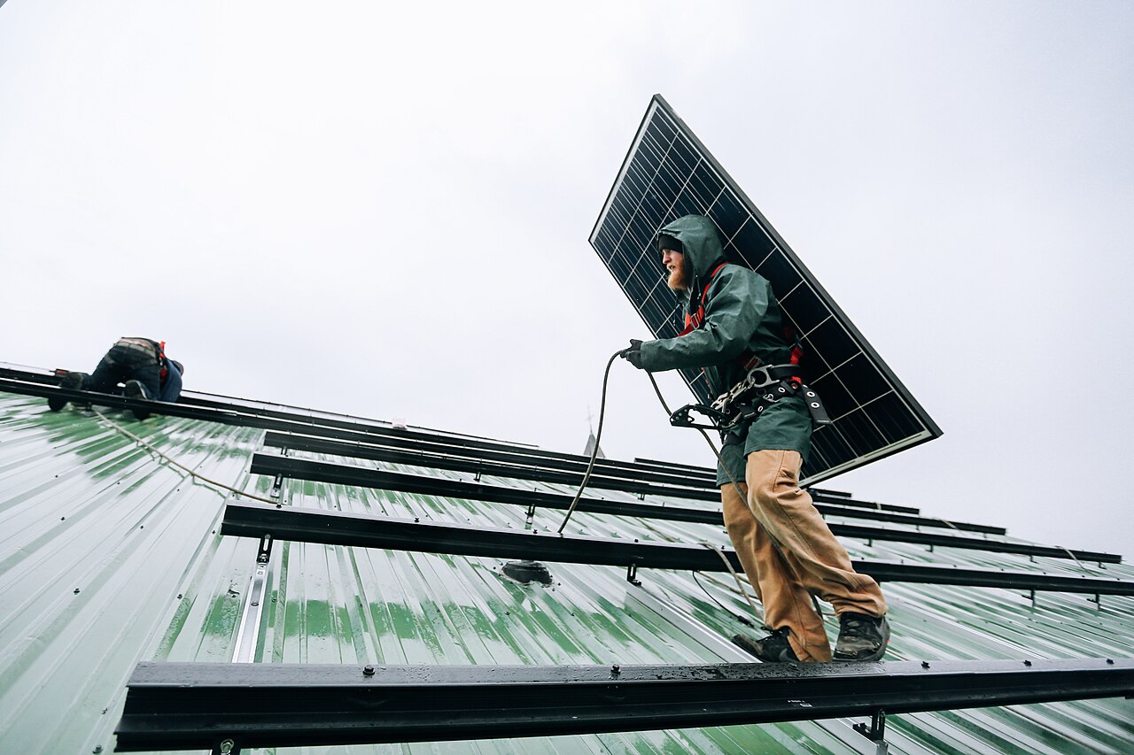 A technician carrying a solar panel across the metal mounting rails bolted to a roof, showing the racking a rooftop solar array is fastened to