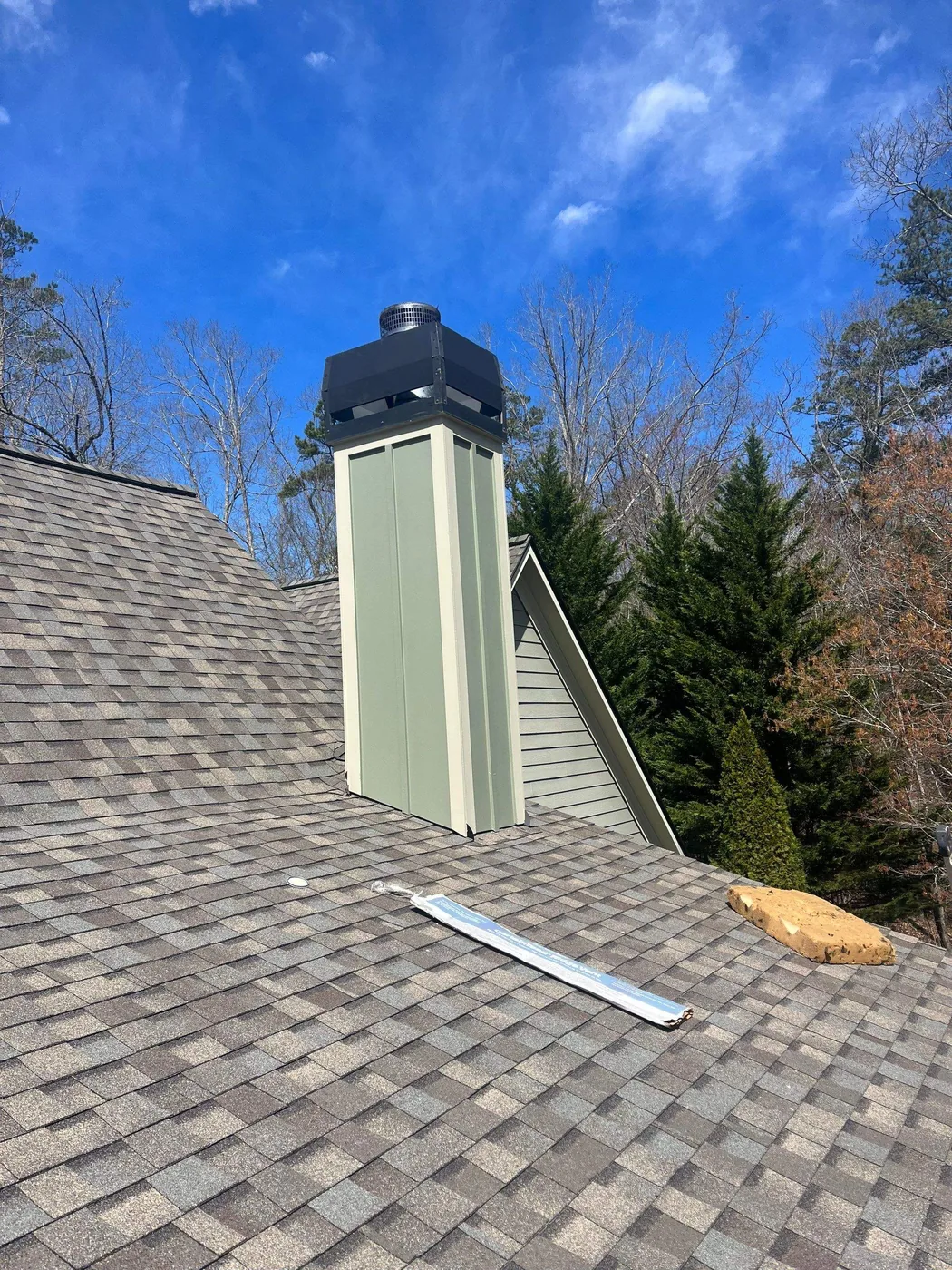 A tall green framed chimney chase on an asphalt shingle roof, topped with a black metal shroud and a screened flue cap