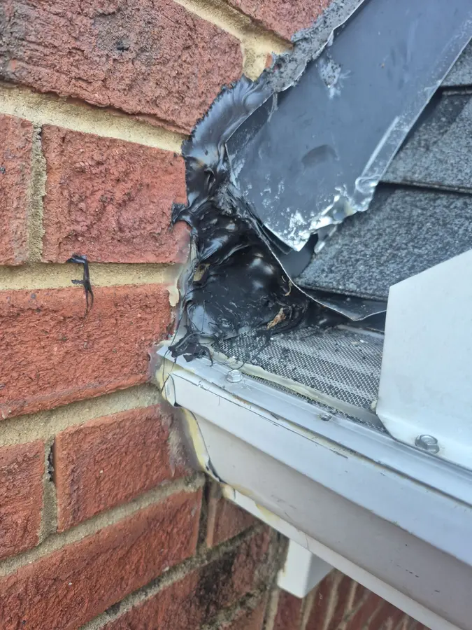 Torn metal flashing covered with a thick layer of black roofing tar where a roof meets a brick wall above a gutter on an Acworth, GA home